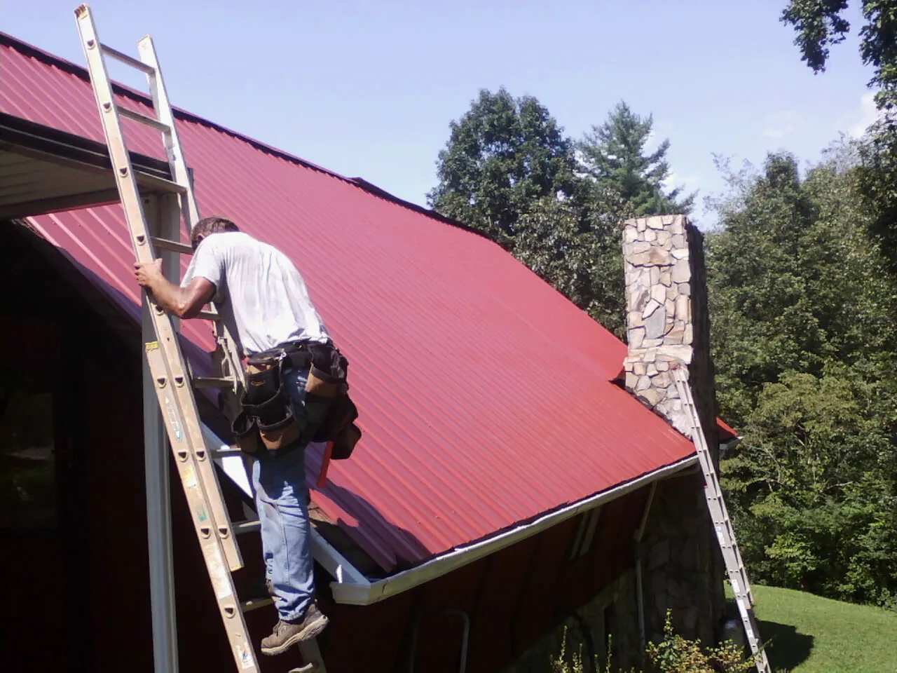 Roofer on a ladder beside a red metal roof and stone chimney — Soffit & Fascia Repair by Roofing Brothers in Glenpool